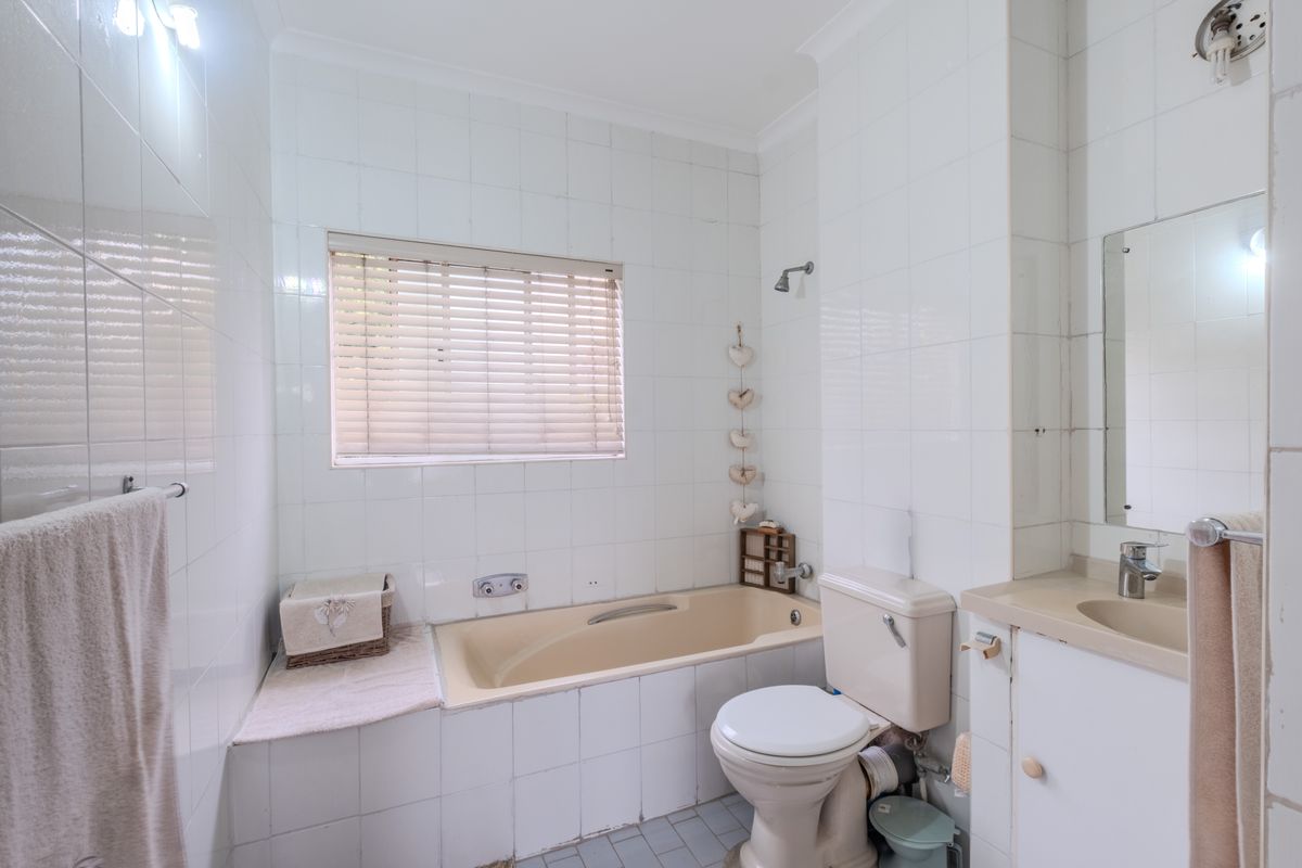 A dated bathroom featuring a built-in bathtub, toilet, and vanity cabinet with integrated sink, finished with white wall tiles and grey floor tiles.