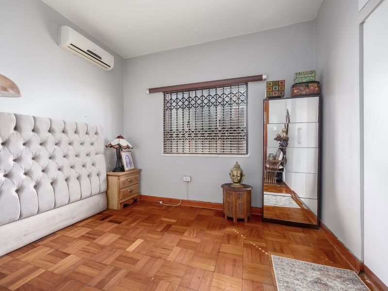 A well-maintained bedroom featuring classic parquet flooring, a wall-mounted air conditioning unit, and a secure window with a fitted security gate.