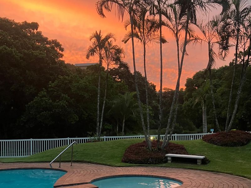 A beautifully landscaped pool and spa area featuring brick paving and a white picket fence, set against a vibrant sunset backdrop.