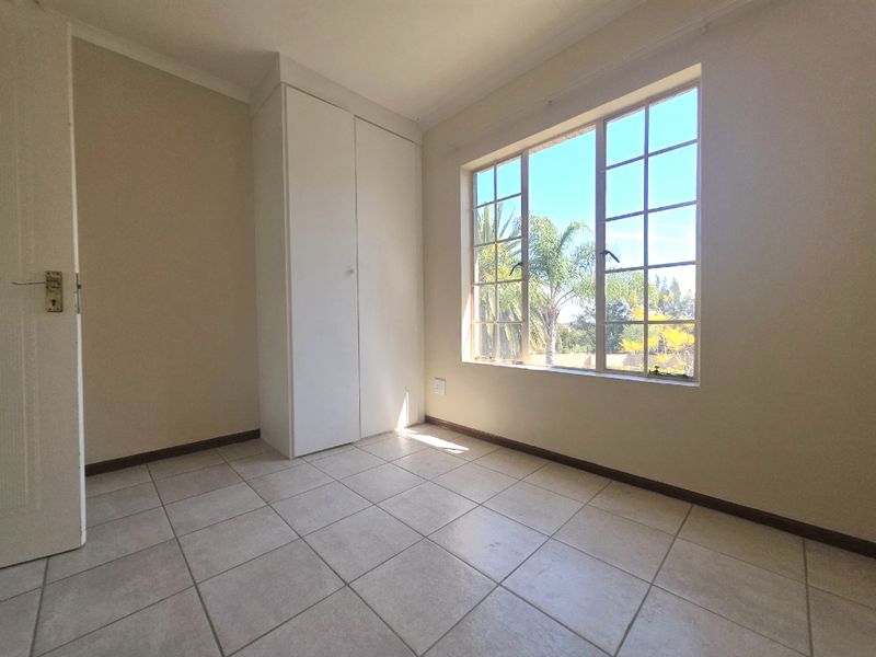 A bright, empty bedroom featuring neutral tiled flooring, a built-in cupboard, and a large window providing natural light and garden views.