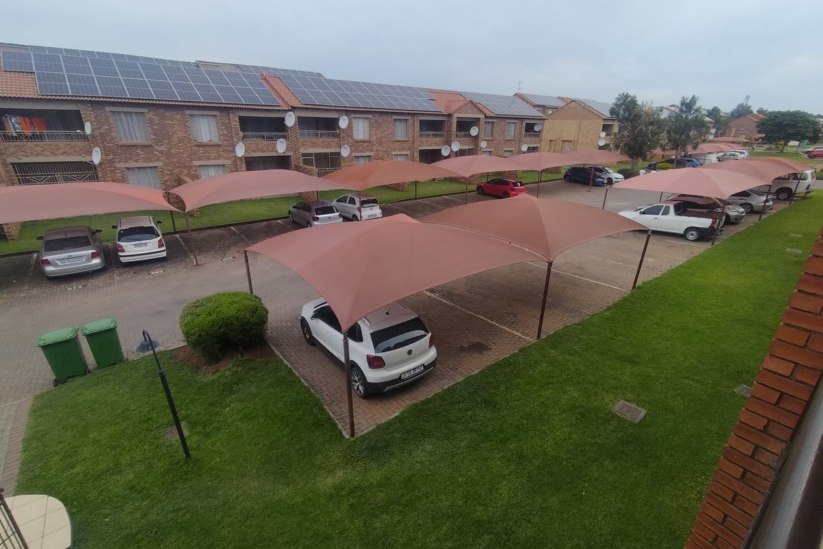 Elevated view of the apartment complex rear exterior, featuring brick building facade, solar panel installation, and covered parking bays with shade cloth structures.