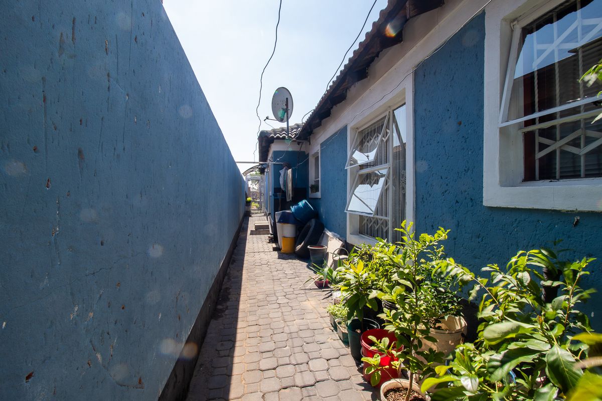 A narrow paved side passage along the exterior of the house featuring blue textured walls, security window bars, and a tiled roof.