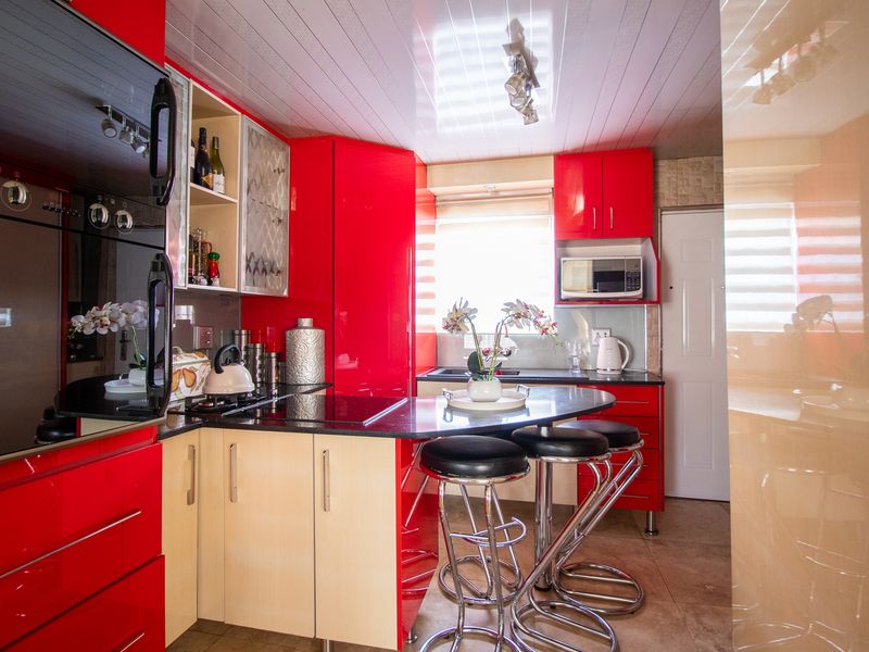 A compact kitchen featuring vibrant red cabinetry, a built-in oven, electric hob, and a functional breakfast bar with stone countertops.