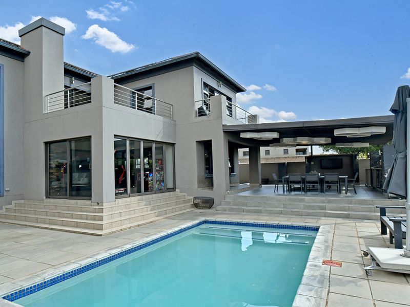 Rear exterior view featuring a swimming pool, tiled deck, covered patio area with built-in bar, and a balcony with stainless steel railings.