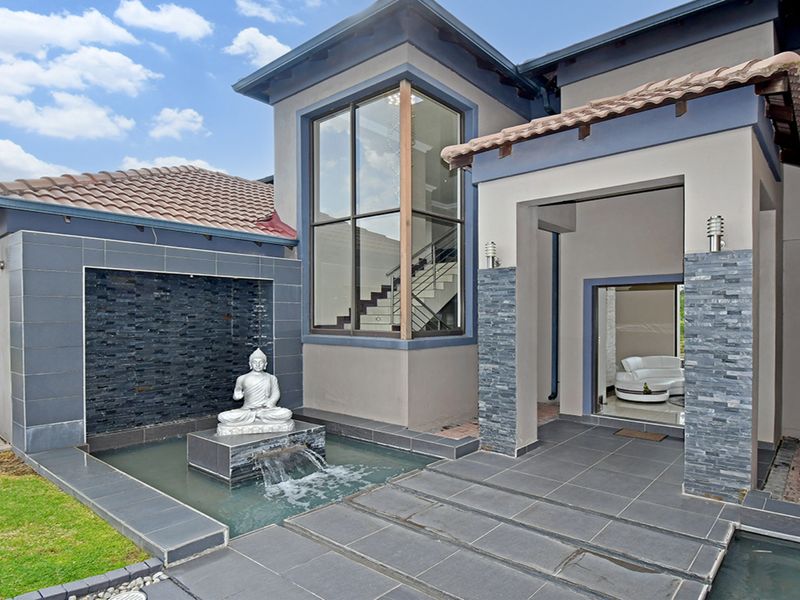 Modern front facade featuring a striking water feature, stone-clad pillars, double-volume windows, and a paved entrance area.
