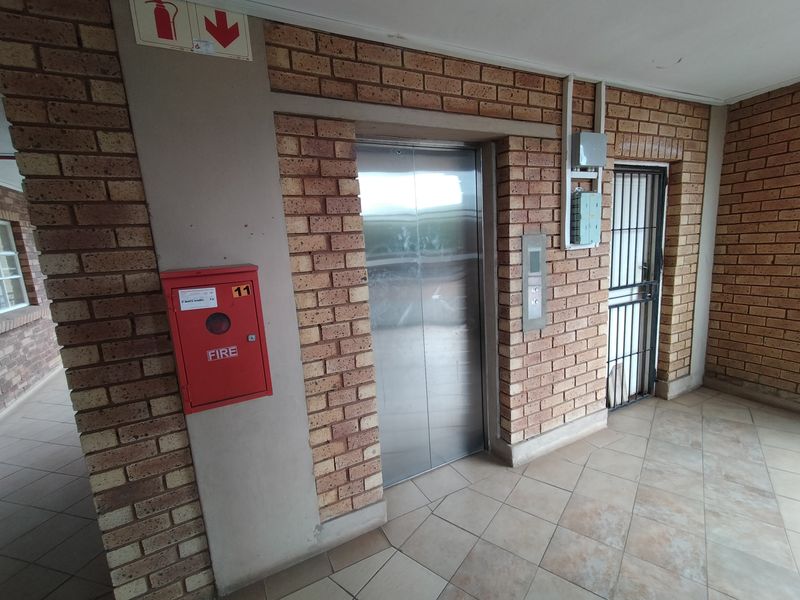 A functional apartment building hallway featuring a stainless steel elevator, fire safety equipment, and a secure entrance gate with exposed brick finishes.