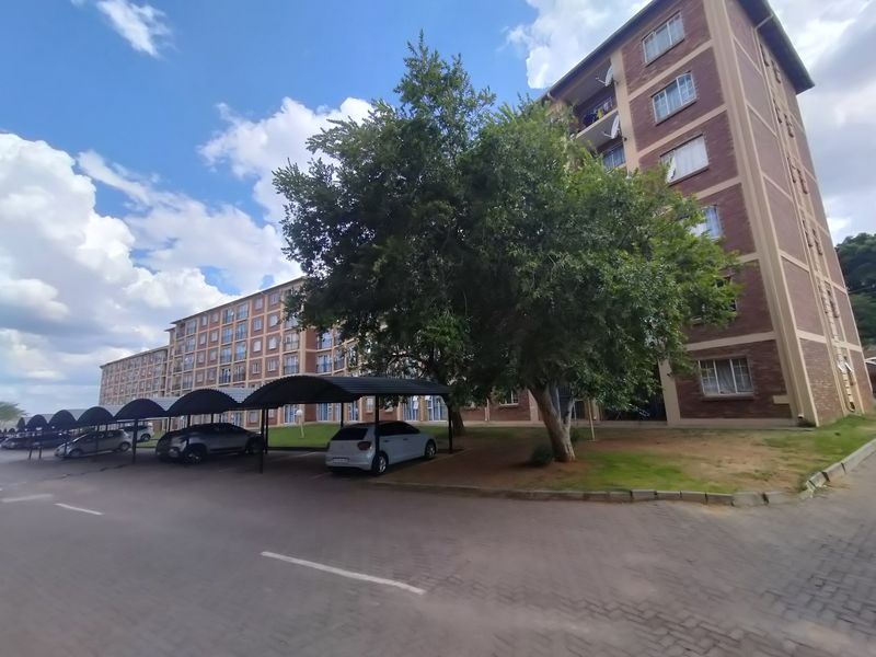 Exterior view of a multi-story brick apartment complex featuring covered parking bays and established trees in the surrounding grounds.