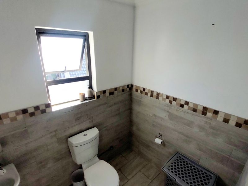 A functional bathroom featuring neutral wood-look wall and floor tiles, a standard toilet, and a washbasin with a top-hung window for natural ventilation.