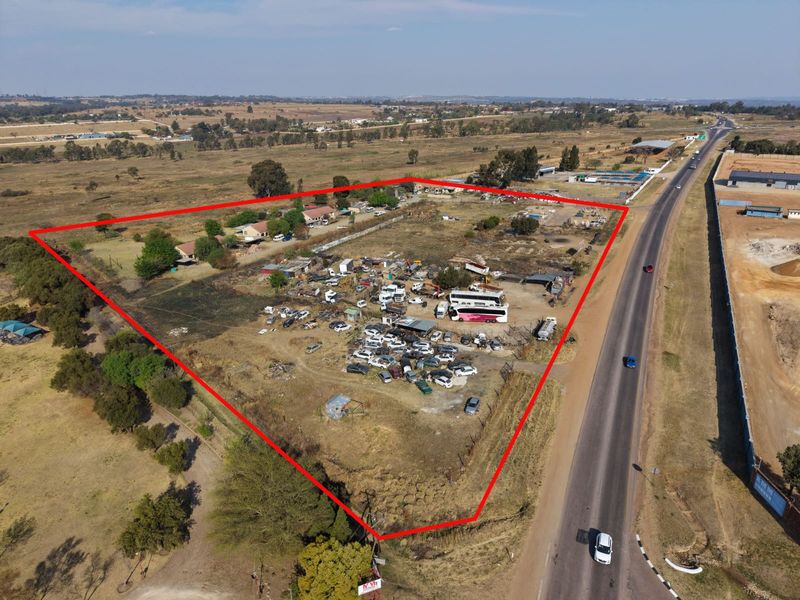 Aerial view of a large, undeveloped plot of land adjacent to a main road, featuring existing outbuildings and significant open space.