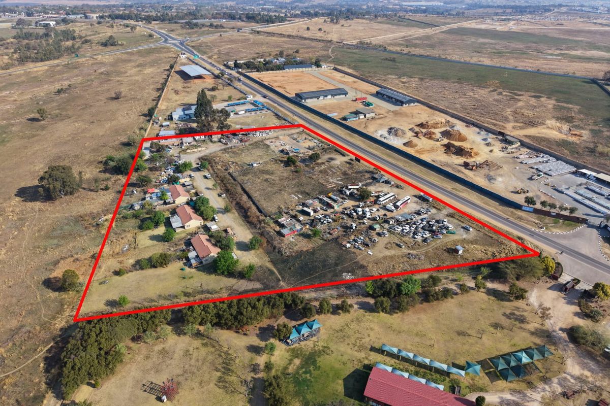 Aerial view of a large, undeveloped plot of land with existing structures, outlined in red, situated near a main road.
