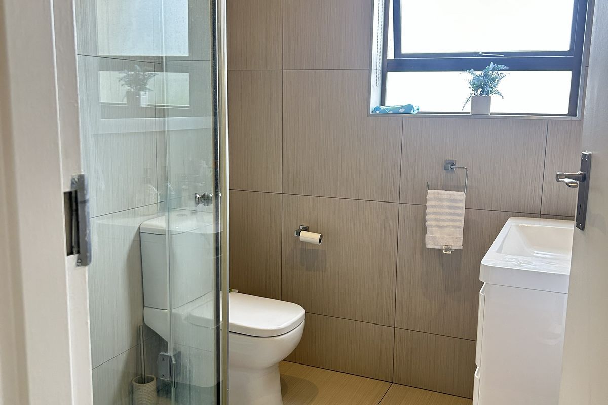 Modern bathroom featuring a glass shower enclosure, wall-mounted vanity, and neutral floor-to-ceiling tiling with a functional top-hung window.