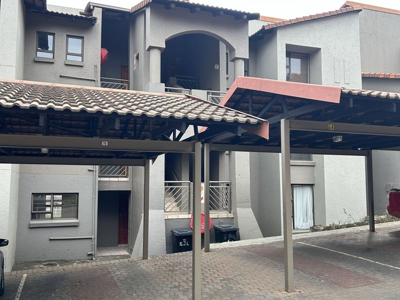 Exterior view of an apartment complex featuring covered parking bays, paved driveway, and multi-level building facade with tiled roofing.