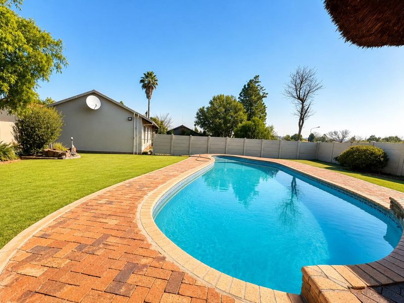 A well-maintained outdoor pool area featuring a curved swimming pool surrounded by brick paving and a manicured lawn with perimeter fencing.