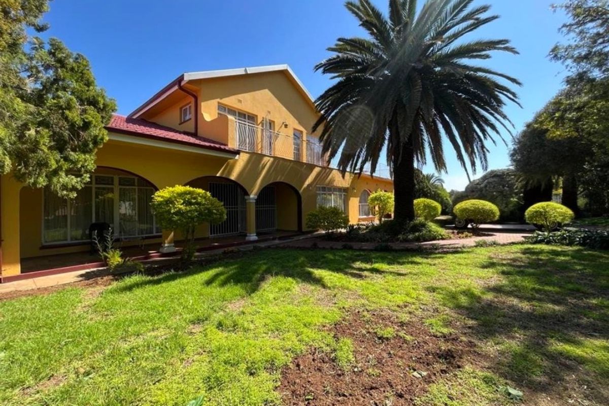 A rear view of a yellow double-story house featuring a covered patio, an upper-level balcony, and a spacious landscaped garden with mature palm trees.