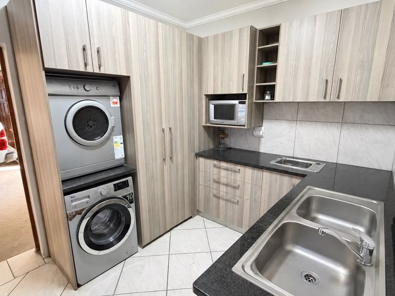 A functional laundry room featuring modern wood-look cabinetry, granite countertops, a double stainless steel sink, and tiled finishes.