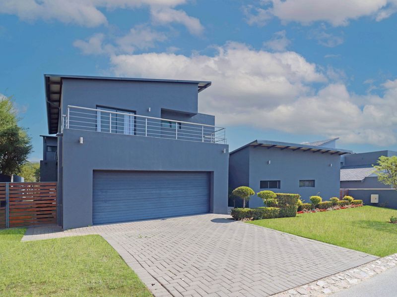 Modern double-story home featuring a paved driveway, double garage, and an upper-level balcony with metal railings, set within a well-maintained landscaped garden.