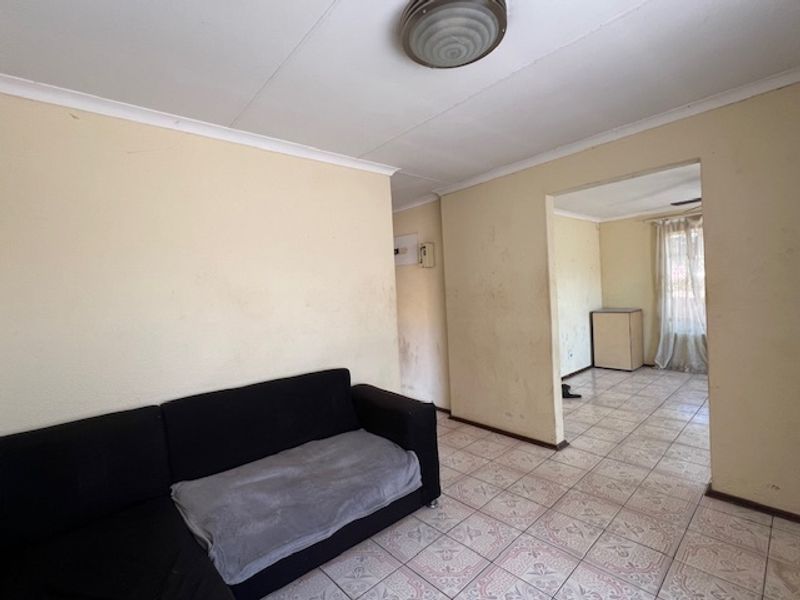 A living area featuring ceramic floor tiles and coved ceilings, leading into an adjacent room. The space requires cosmetic updates to the walls and flooring.