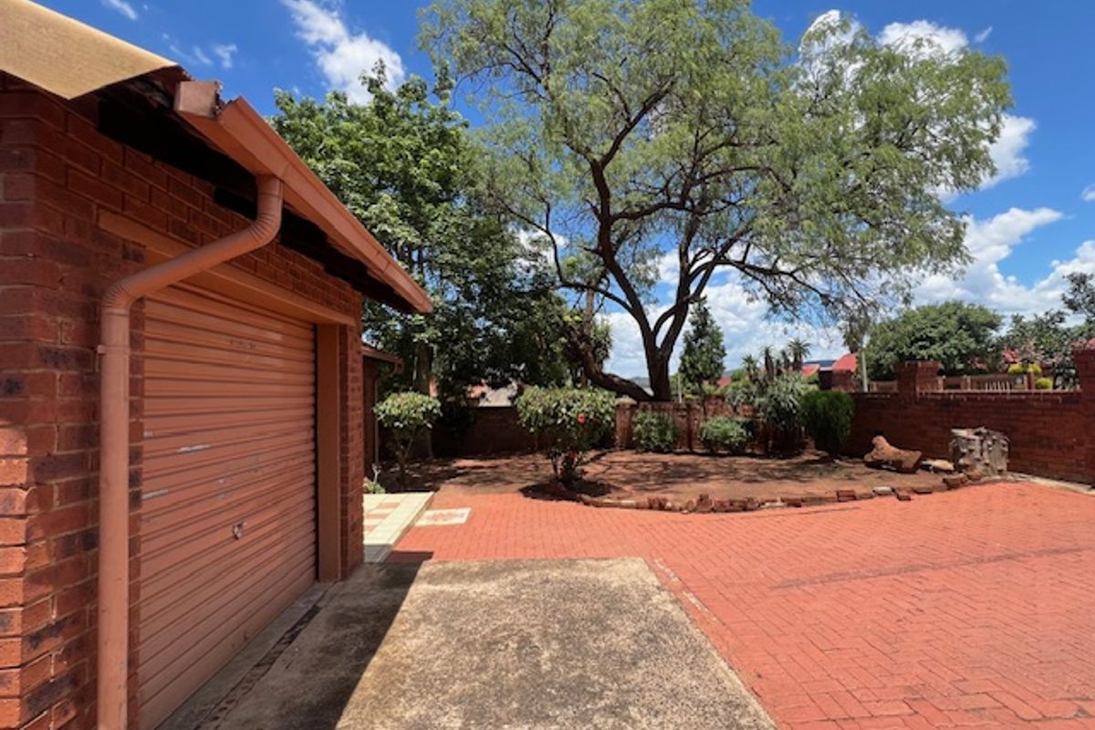 Rear exterior view featuring a brick garage with a roller door, paved driveway, and established garden area with boundary walling.