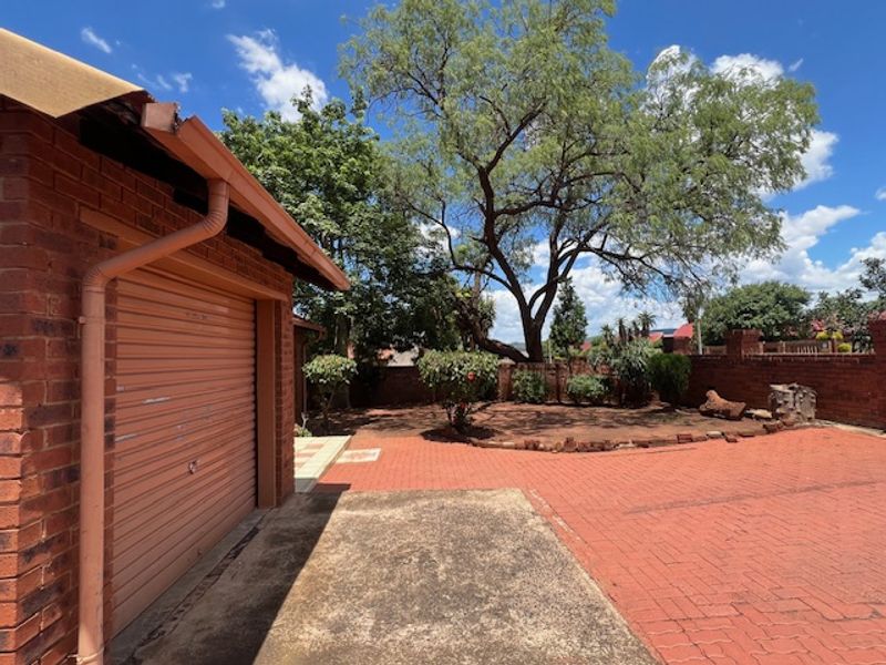 Rear exterior view featuring a brick garage with a roller door, paved driveway, and established garden area with boundary walling.
