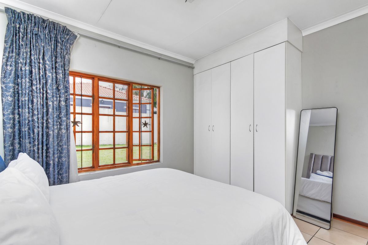 A bedroom featuring built-in white cupboards, tiled flooring, and wooden window frames overlooking the garden.