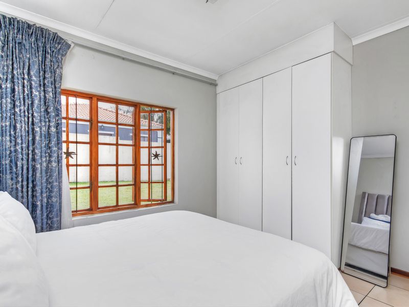 A bedroom featuring built-in white cupboards, tiled flooring, and wooden window frames overlooking the garden.