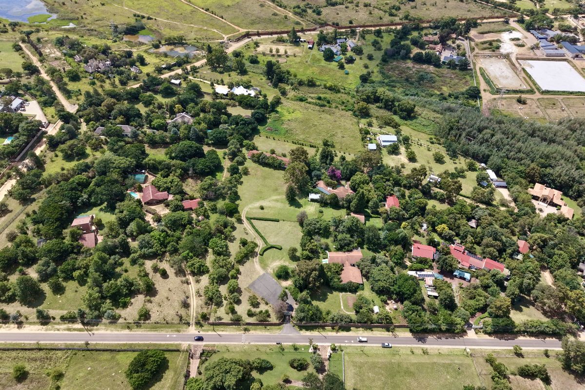 An aerial view of a residential property featuring expansive green grounds, mature trees, and established perimeter fencing in a semi-rural setting.