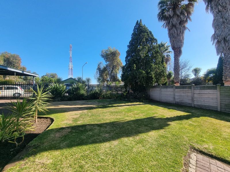 A spacious private garden featuring a well-maintained lawn, mature palm trees, and secure perimeter walling with a paved driveway area.