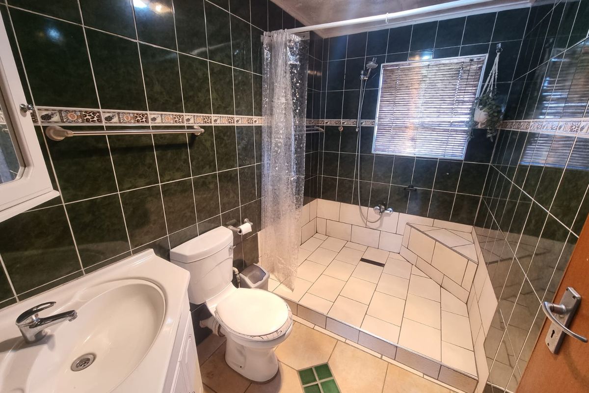 A functional bathroom featuring a walk-in shower with tiled surround, vanity basin, and toilet, finished with dark green wall tiles and neutral floor tiles.