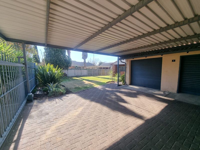A covered carport area featuring a paved driveway, roller shutter garage door, and secure metal fencing, leading to a private garden space.