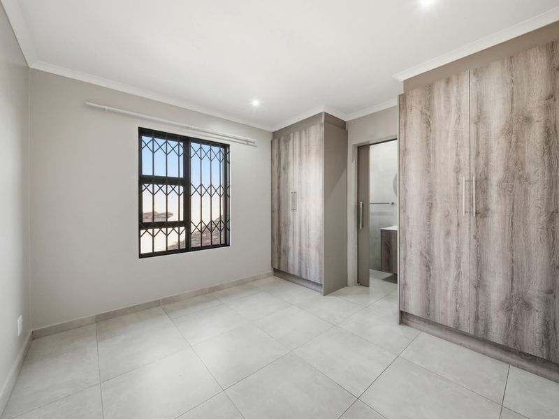A bright, modern bedroom featuring neutral tiled flooring, built-in wood-grain cupboards, and a window fitted with security bars.