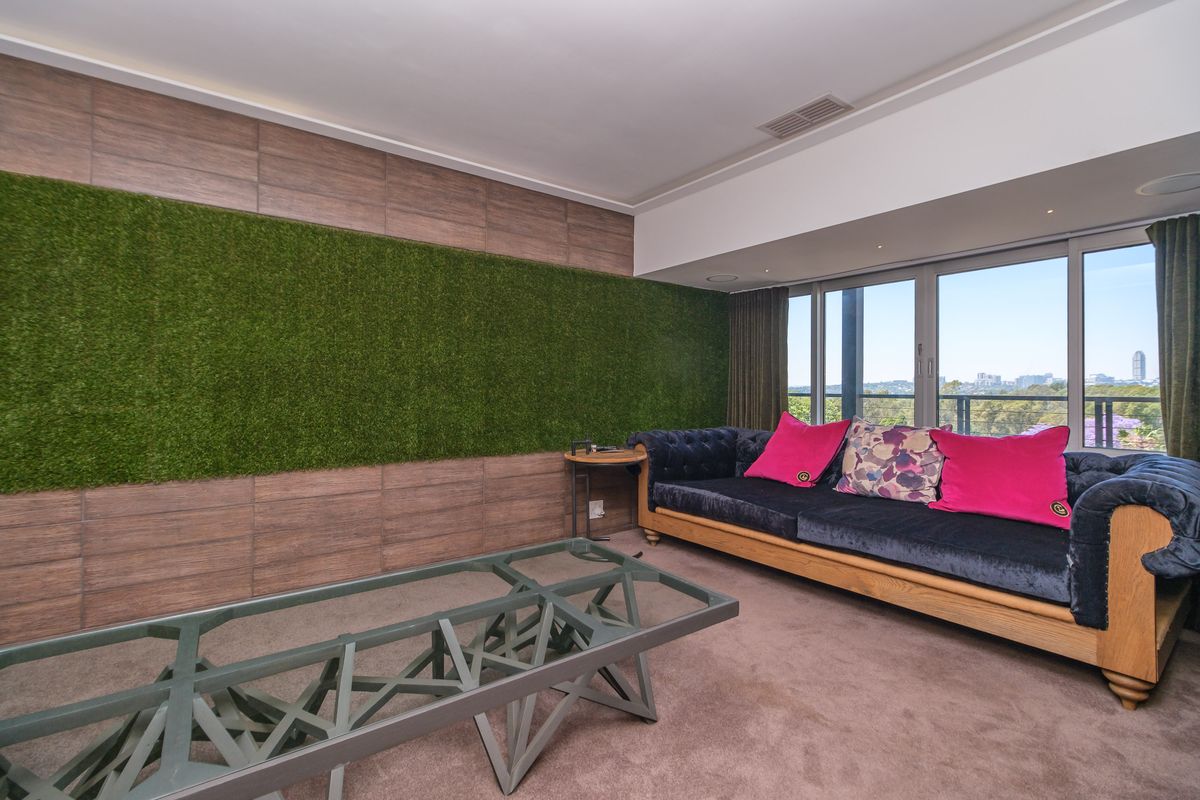 A modern living room featuring a unique artificial grass feature wall, recessed ceiling lighting, and large sliding glass doors offering expansive city views.