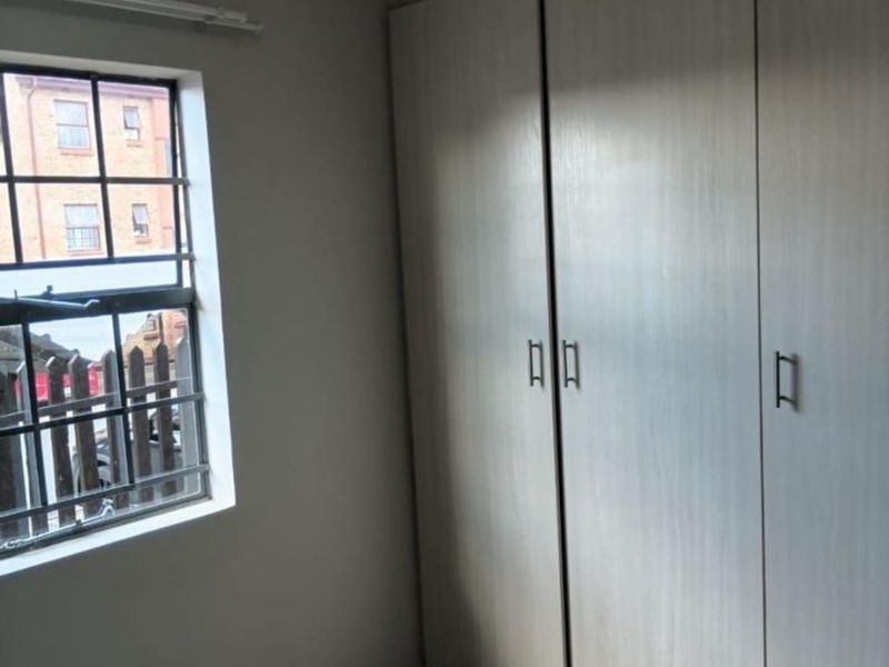 A clean, empty bedroom featuring neutral tiled flooring and ample built-in cupboard storage, with a window fitted with security bars.