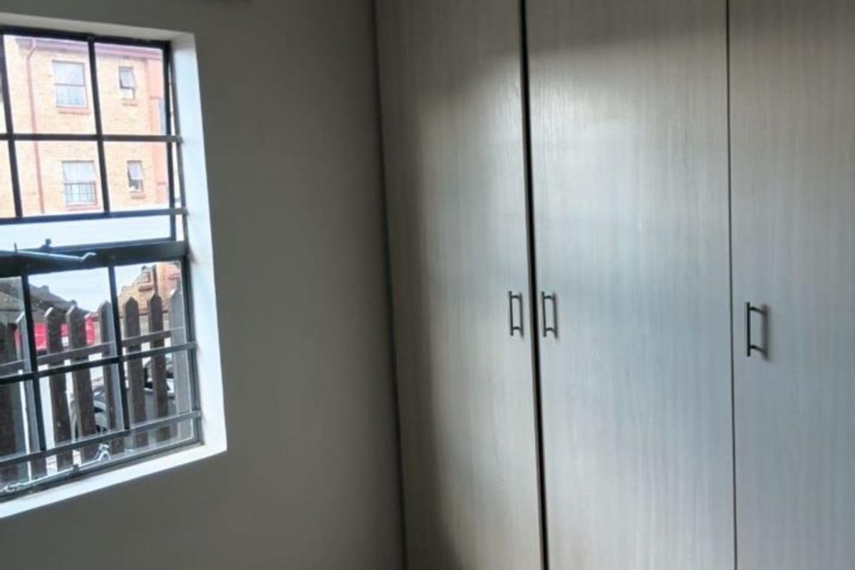 A clean, empty bedroom featuring neutral tiled flooring and ample built-in cupboard storage, with a window fitted with security bars.