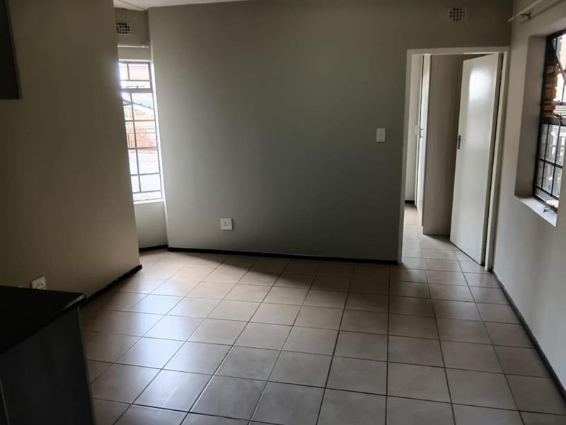 An empty, neutrally painted living area featuring tiled flooring, natural light from multiple windows, and direct access to an adjacent hallway.