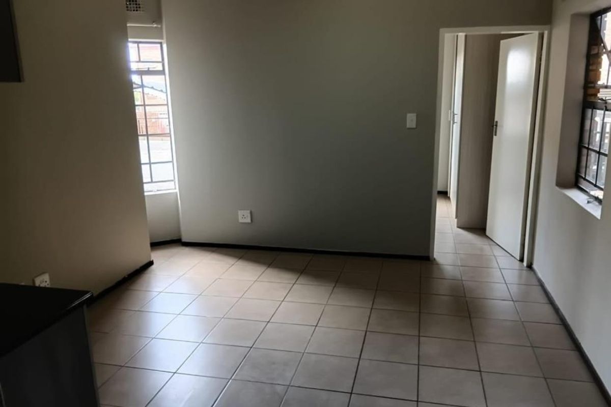 An empty, neutrally painted living area featuring tiled flooring, natural light from multiple windows, and direct access to an adjacent hallway.
