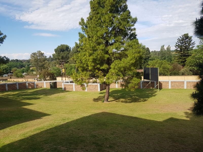 A spacious communal garden area featuring a well-maintained lawn, mature trees, and a secure brick perimeter wall.