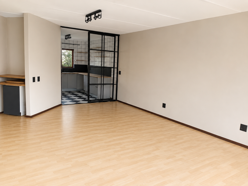 An open-plan living area featuring light laminate flooring, neutral walls, and a stylish black metal-framed glass partition leading into a modern kitchen.