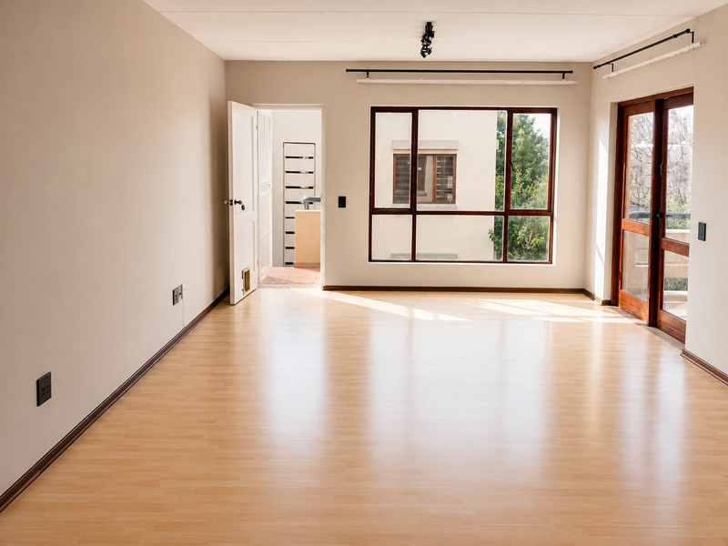 A bright, empty living room featuring light-toned laminate flooring, large windows, and direct access to an outdoor area through a glass panel door.