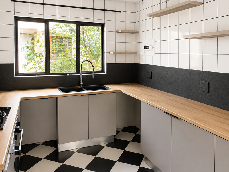 Modern kitchen featuring light grey cabinetry, wooden countertops, black double sink, and a striking checkered floor with white tiled backsplash and floating shelves.