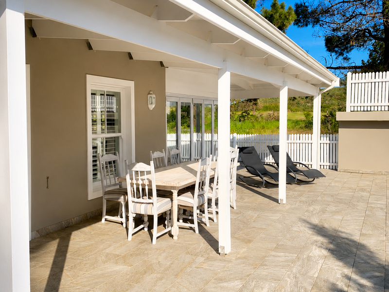 A spacious covered patio featuring tiled flooring, timber support posts, and folding glass doors, overlooking a landscaped garden with a white picket fence.