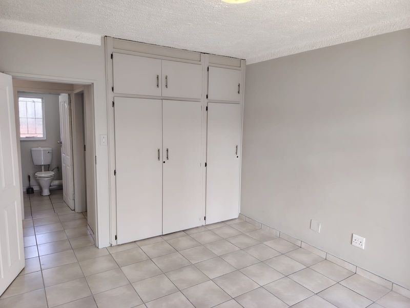 A clean, empty bedroom featuring neutral tiled flooring, built-in cupboards, and a textured ceiling, with an adjacent view into a bathroom.