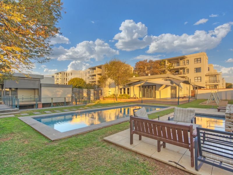 A well-maintained outdoor pool area featuring a swimming pool, paved decking, and landscaped garden space within a residential apartment complex.