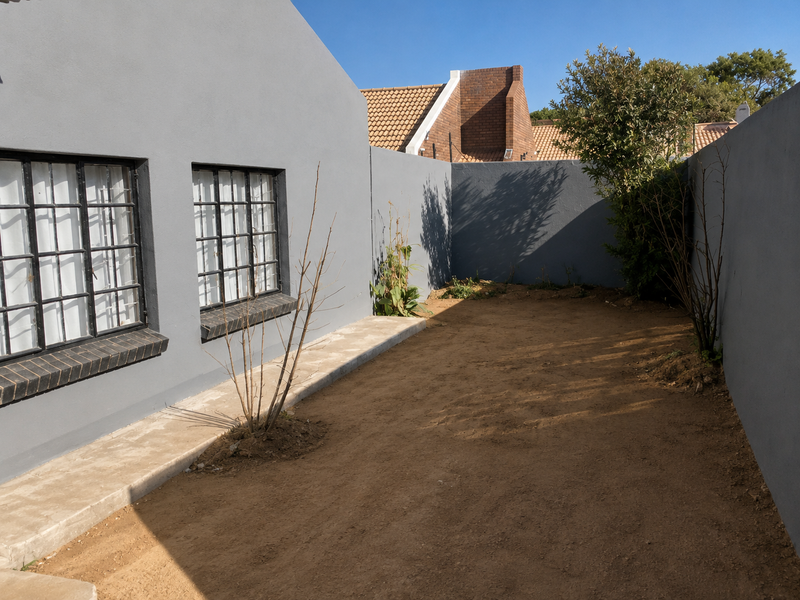 A private rear courtyard area featuring grey rendered walls, steel-framed windows, and a concrete pathway, currently awaiting landscaping.