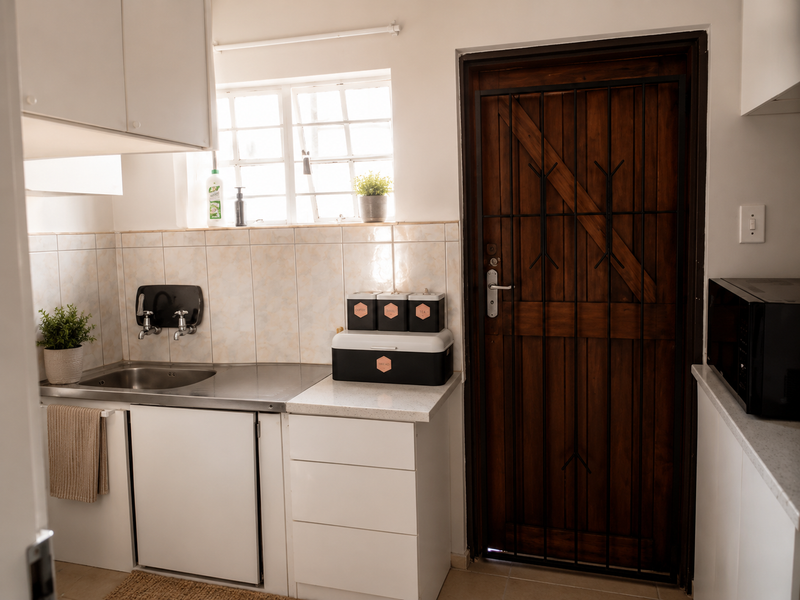 A functional scullery area featuring white cabinetry, a stainless steel sink, tiled splashback, and a solid timber door with a security gate.