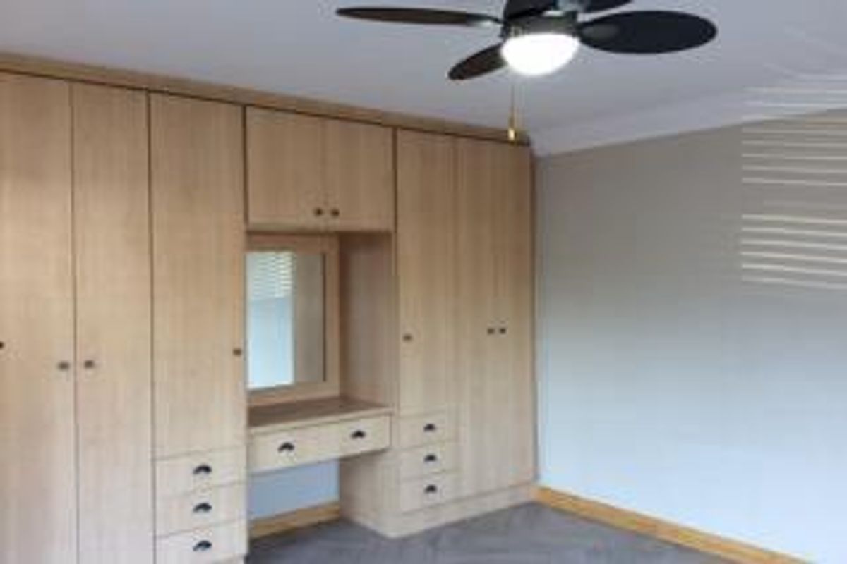 A bedroom featuring extensive light wood built-in cupboards with an integrated vanity area and a ceiling fan.