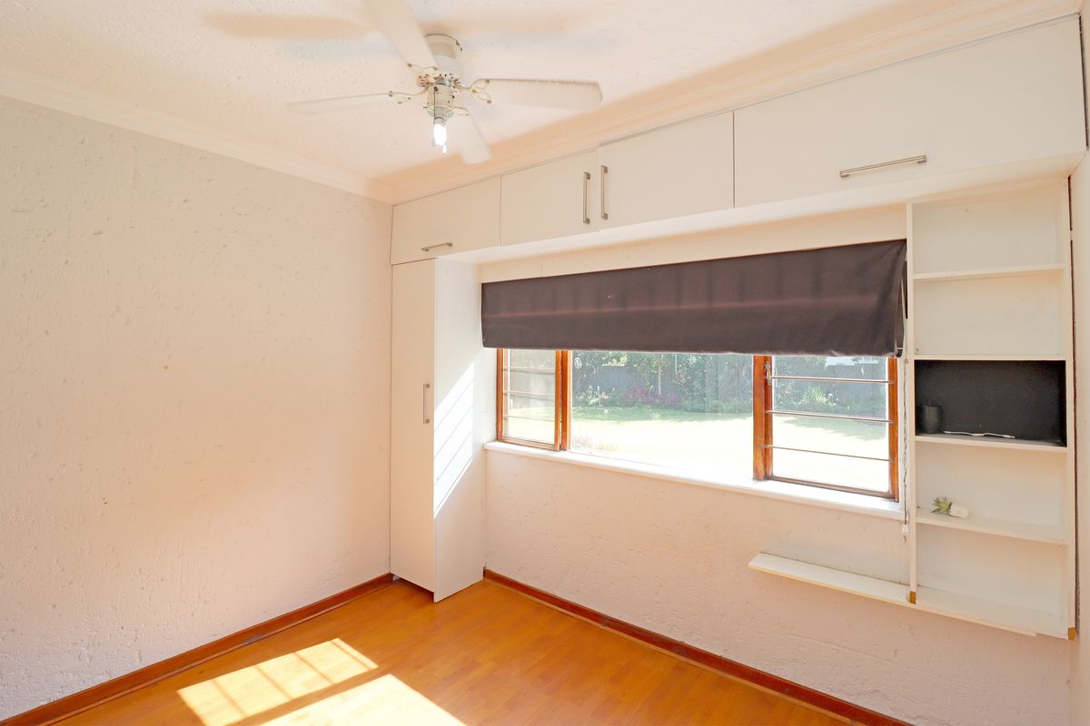 A bright, empty bedroom featuring built-in cupboards, integrated shelving, wooden flooring, and a ceiling fan, overlooking the garden.