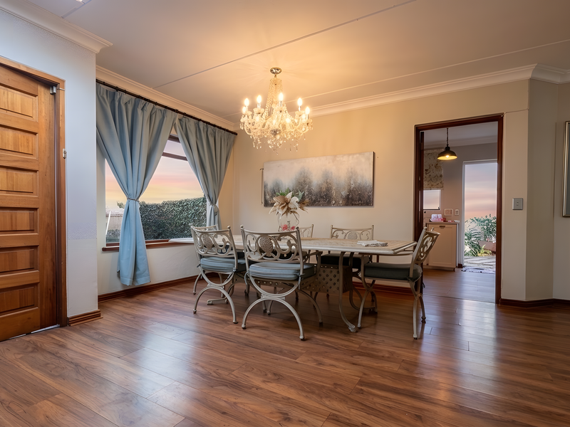 A well-maintained dining room featuring warm laminate flooring, a classic crystal chandelier, and a solid timber door, with a view into the adjacent kitchen area.