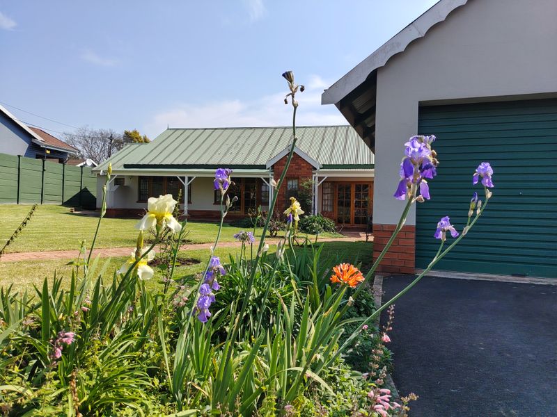 A well-maintained rear exterior view featuring a brick house with a green corrugated metal roof, a paved driveway, and a landscaped garden.
