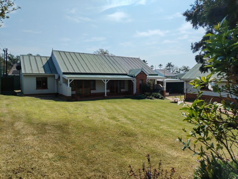 A well-maintained rear exterior view of the house featuring a large lawn, covered patio area, and traditional brick and metal roof construction.