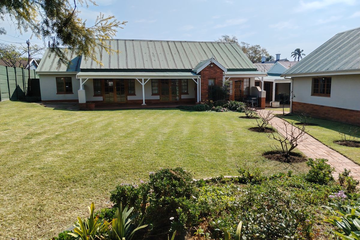 A well-maintained rear exterior view of the house featuring a spacious lawn, brick paved pathways, and a covered patio area with timber framed doors.